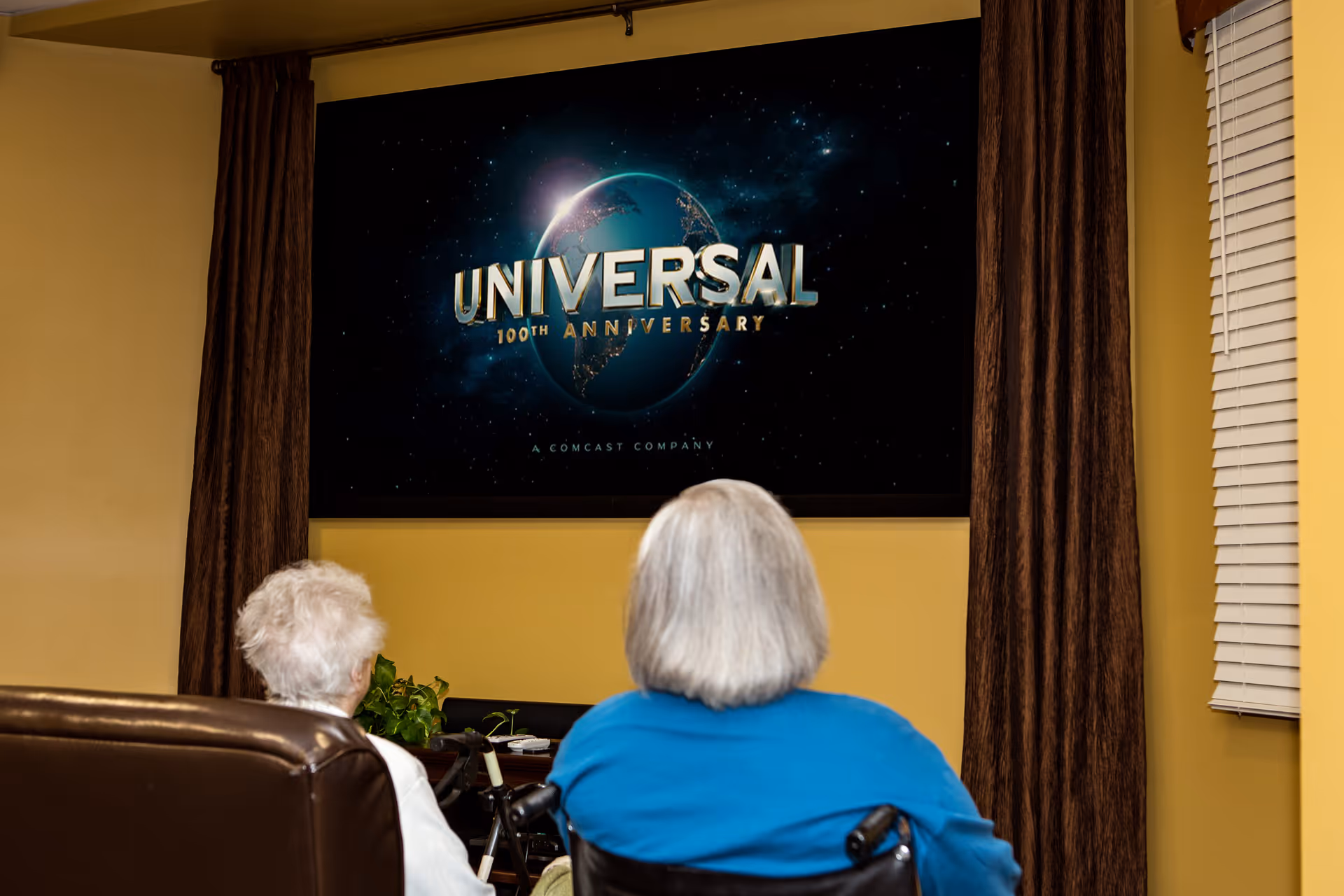 Two elderly individuals sitting in a living room area watching a Universal 100th Anniversary movie on a large wall-mounted TV screen. One person is seated in a wheelchair and the other in a brown leather chair. The room has yellow walls, brown curtains, and a window with white blinds.