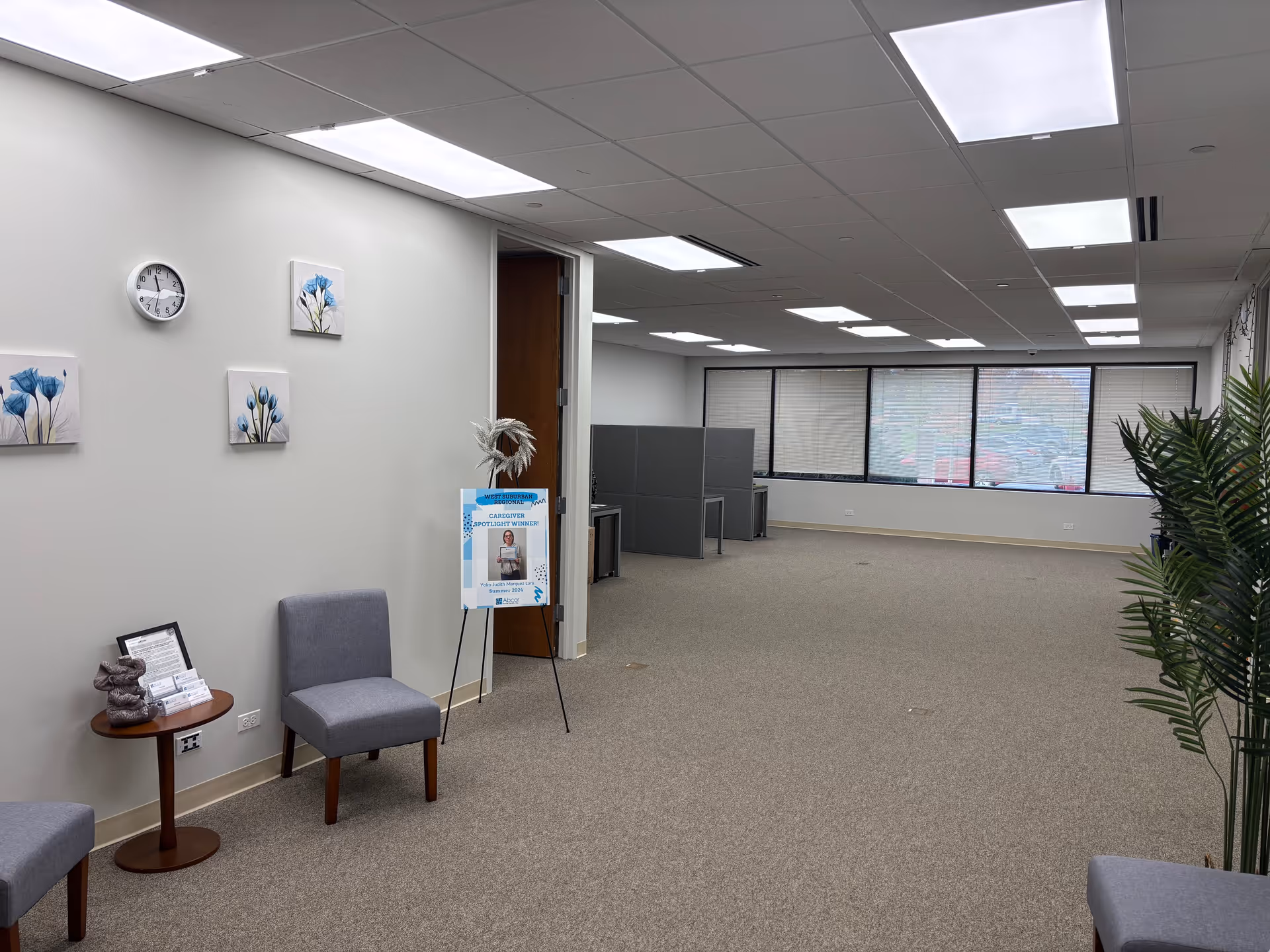 Wide interior lobby with grey chairs, wall art, clock, signboard, and large windows.