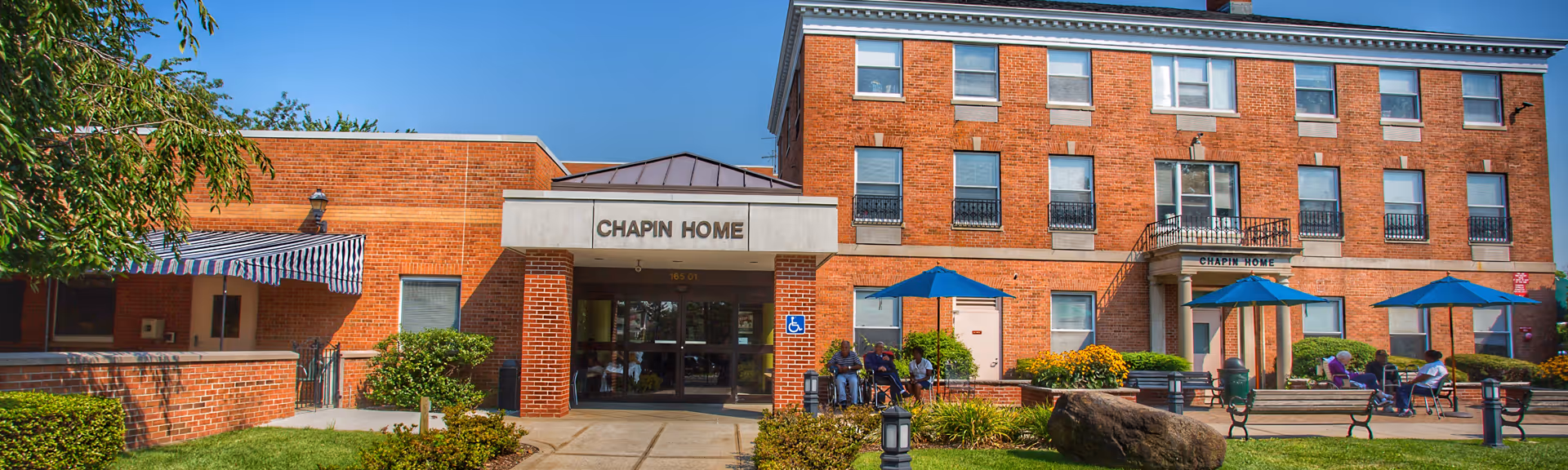 Exterior view of Chapin Home for the Aging, a multi-story brick building with a main entrance canopy. There are benches, blue umbrellas, and people sitting outside on a sunny day with a clear blue sky.