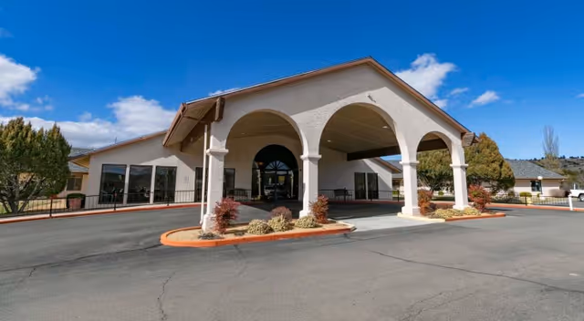 Front entrance of a single-story senior living facility with a covered porte-cochère supported by arched columns under a blue sky.