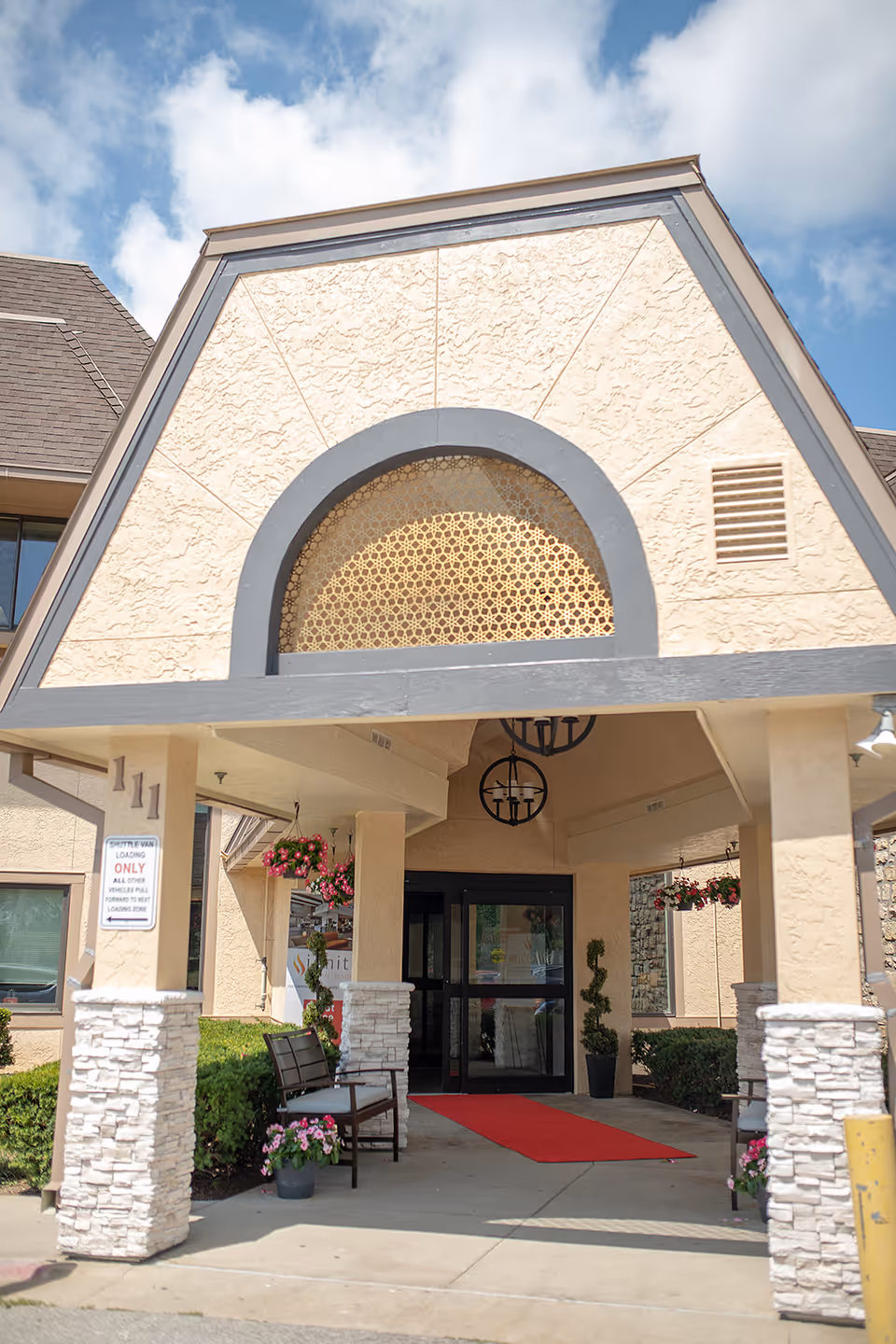 Entrance of Luxe Life Senior Living and Memory Care St. Mary's with a covered driveway, stone pillars, hanging flower baskets, benches, and a red carpet leading to glass doors under a decorative archway.