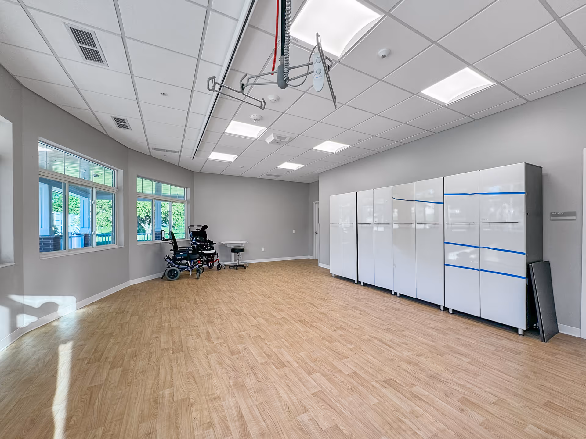A spacious room with light wood flooring and gray walls, featuring large windows on one side letting in natural light. There are several white storage cabinets lined up against one wall, two wheelchairs near the windows, and a ceiling-mounted patient lift system.
