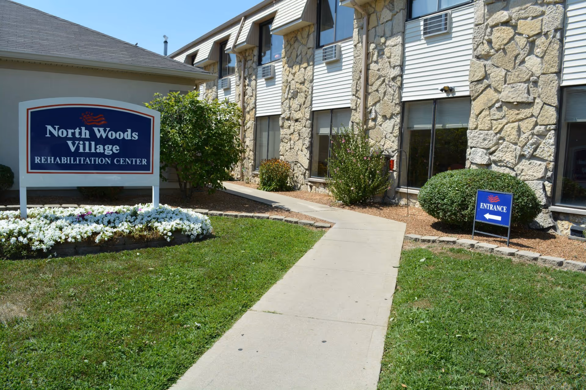 Front exterior of the North Woods Village rehabilitation building with a sign, walkway, and landscaped flower beds.