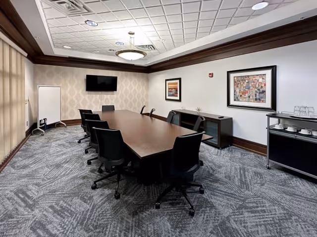 A conference room with a long wooden table, black chairs, a wall TV, and framed art.