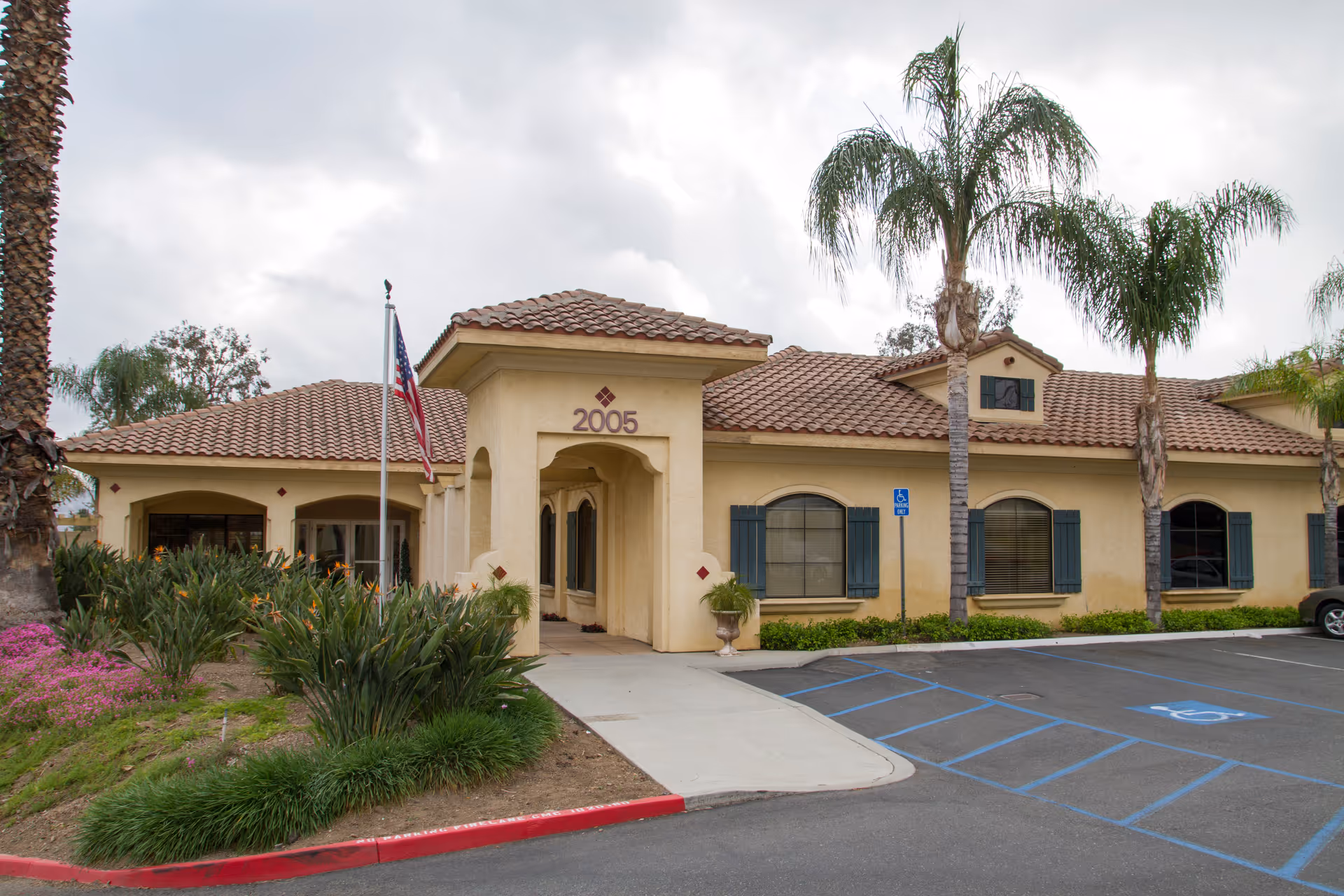 Beige senior living facility exterior with palm trees, a tiled roof, and an American flag.