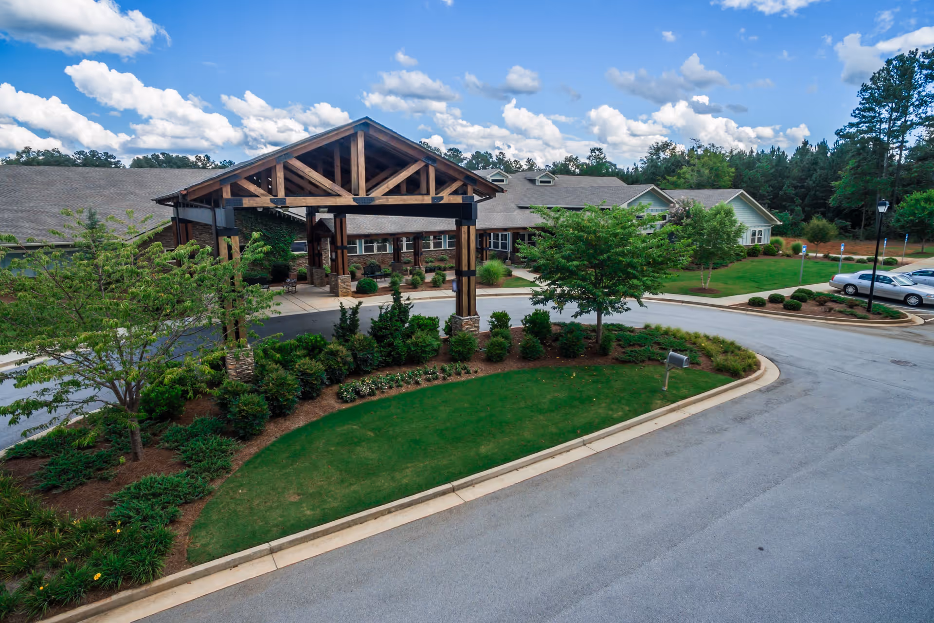 Exterior view of a senior living facility with a large covered entrance supported by wooden beams, surrounded by landscaped greenery and trees under a partly cloudy blue sky.