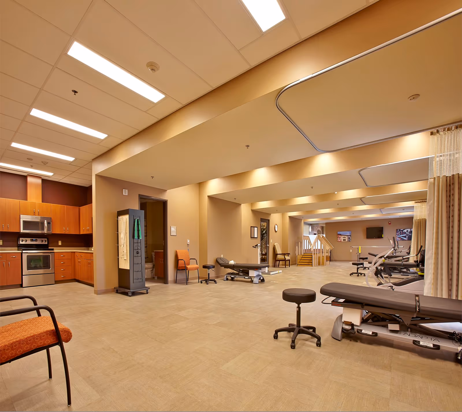 A spacious interior therapy and fitness area with treatment tables, exercise equipment, chairs, and a kitchenette along one wall.