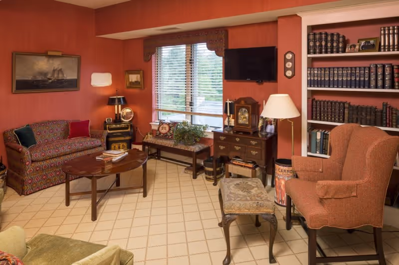 A cozy living room with red walls and beige carpet featuring a patterned sofa with colorful pillows, a wooden coffee table with books, a cushioned bench under a window with blinds, a wooden chest with a vintage clock and lamp, a flat-screen TV mounted on the wall, a tall floor lamp, an upholstered armchair, a small cushioned stool, and a built-in bookshelf filled with books and framed photos.