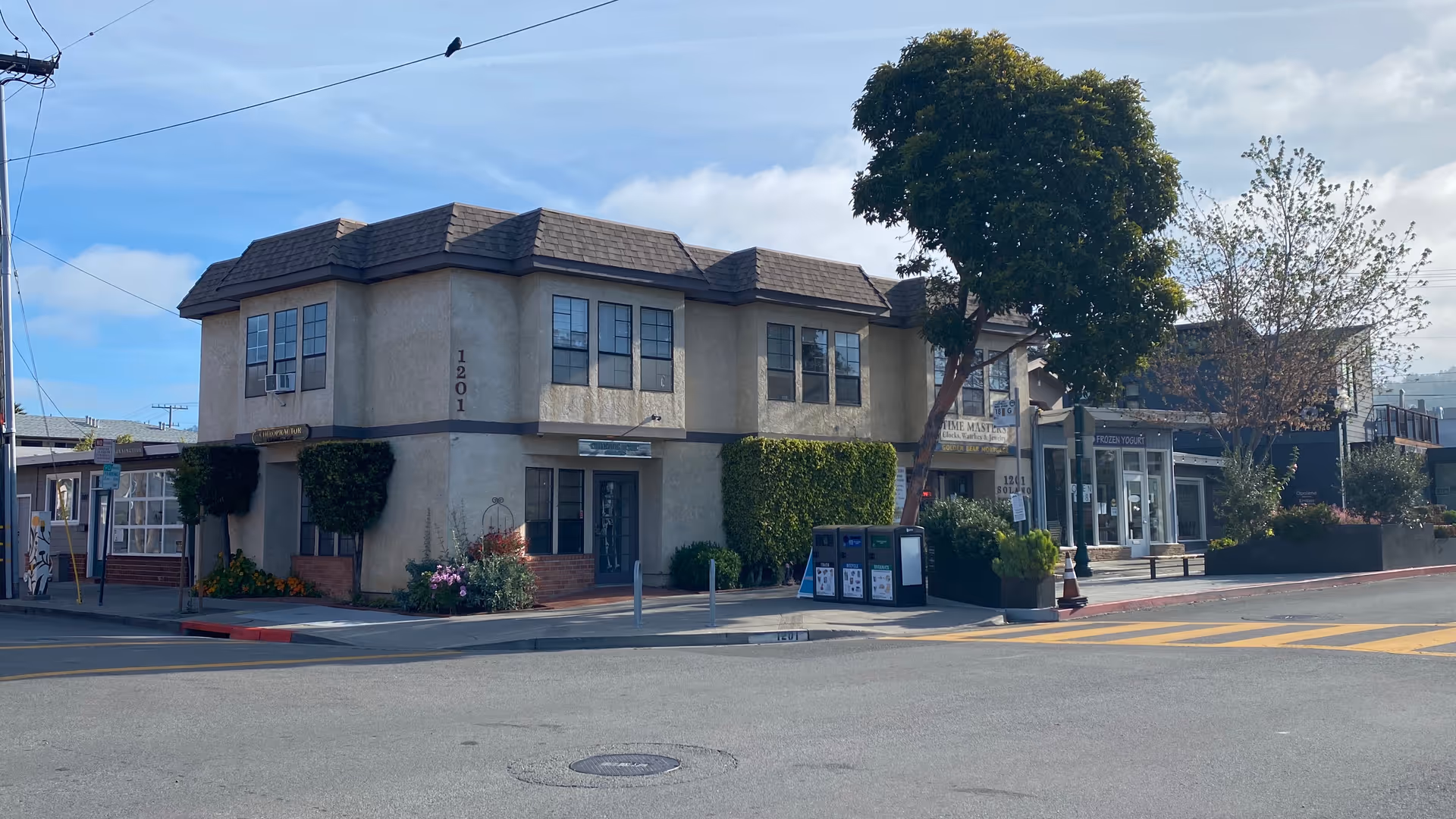 Street view of a two-story beige building with storefronts along a crosswalk.