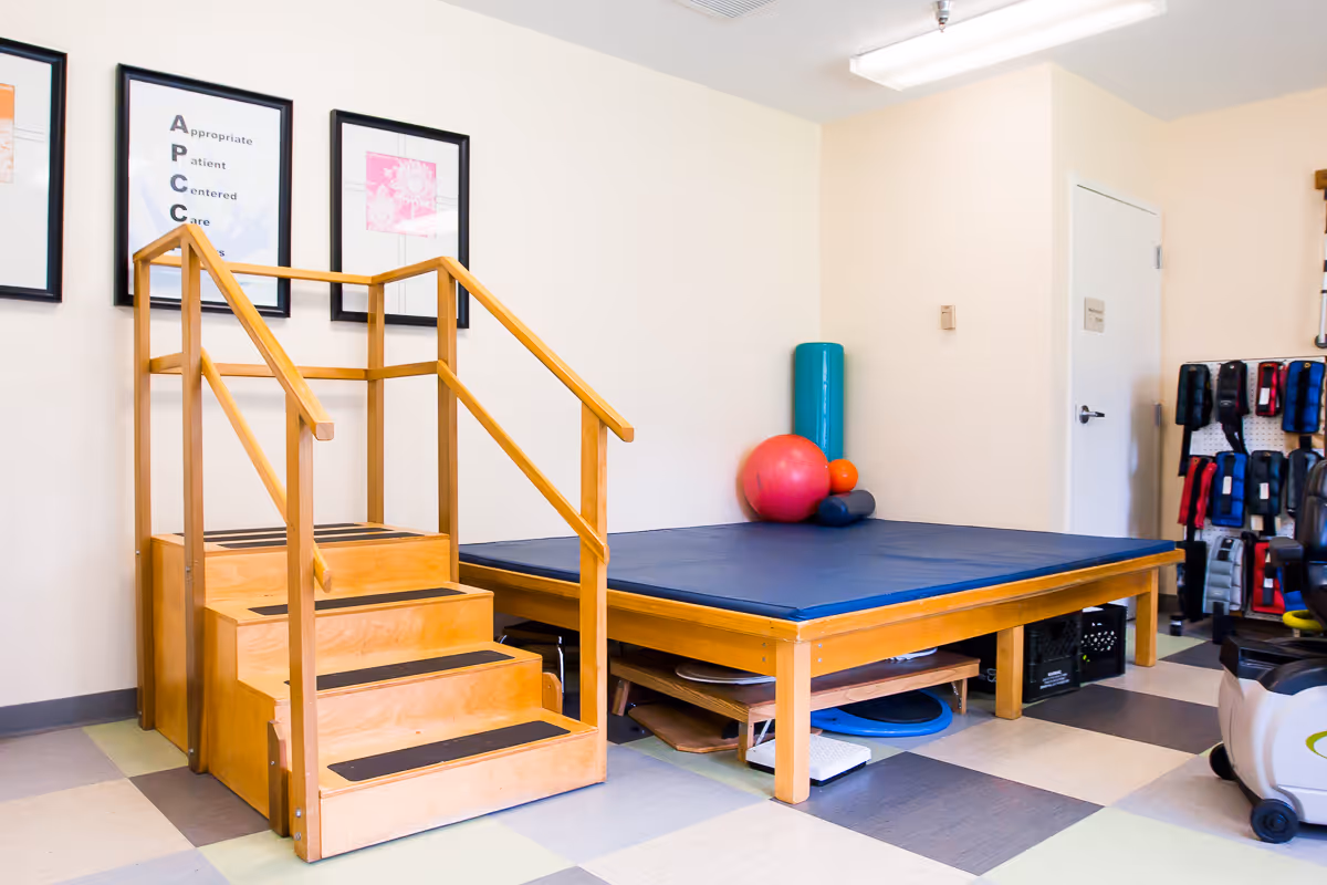 Physical therapy room with wooden practice stairs, a padded exercise platform, therapy balls and assorted equipment.