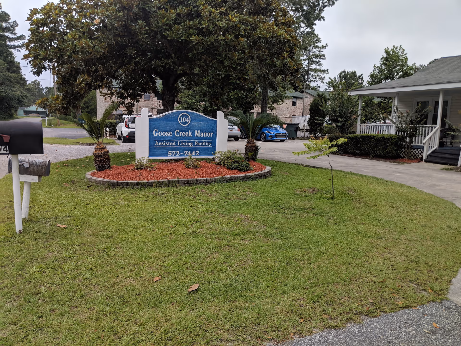 Outdoor view of Goose Creek Manor assisted living facility sign surrounded by grass, small palm trees, and a tree, with a driveway and parked cars in the background.