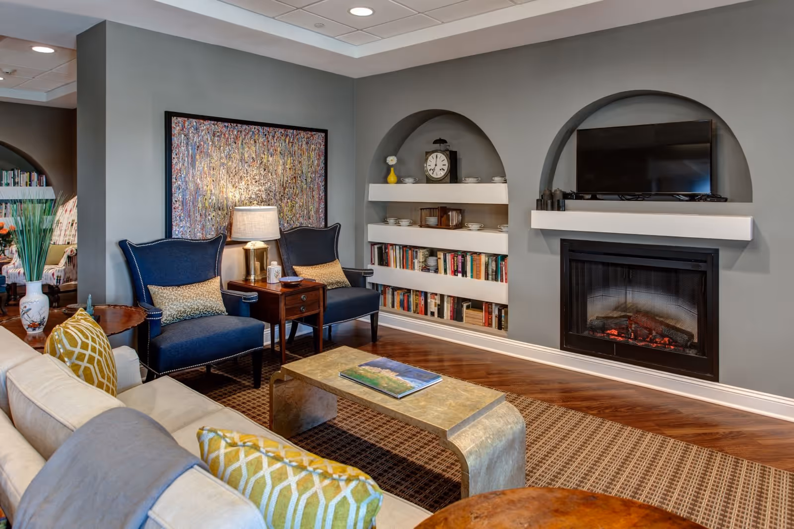 A cozy living room with a beige sofa adorned with yellow and gray pillows, two dark blue armchairs with patterned cushions, a wooden side table with a lamp, a metallic coffee table with a book on it, and built-in shelves with books, dishes, and a clock. There is a fireplace with a TV mounted above it, and a large colorful abstract painting on the wall.