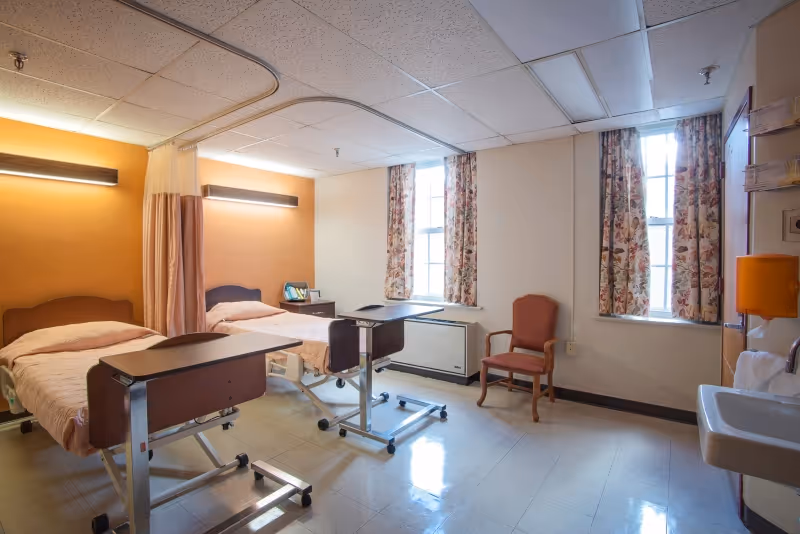 A bright shared patient room with two twin hospital beds, adjustable overbed tables, a chair, sink, and windows with floral curtains.