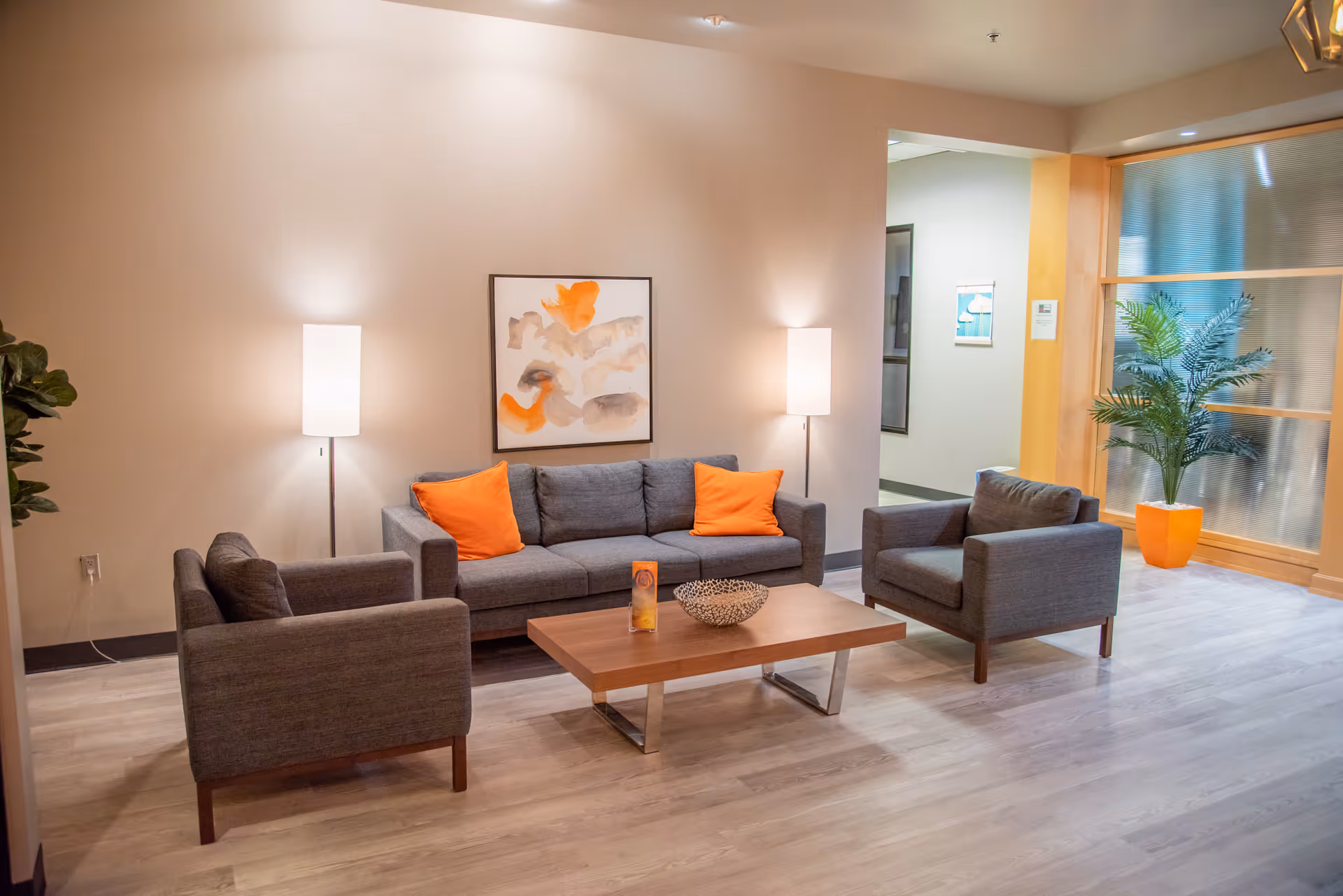 Modern waiting lounge with gray furniture, orange cushions, coffee table, lamps, and plants.