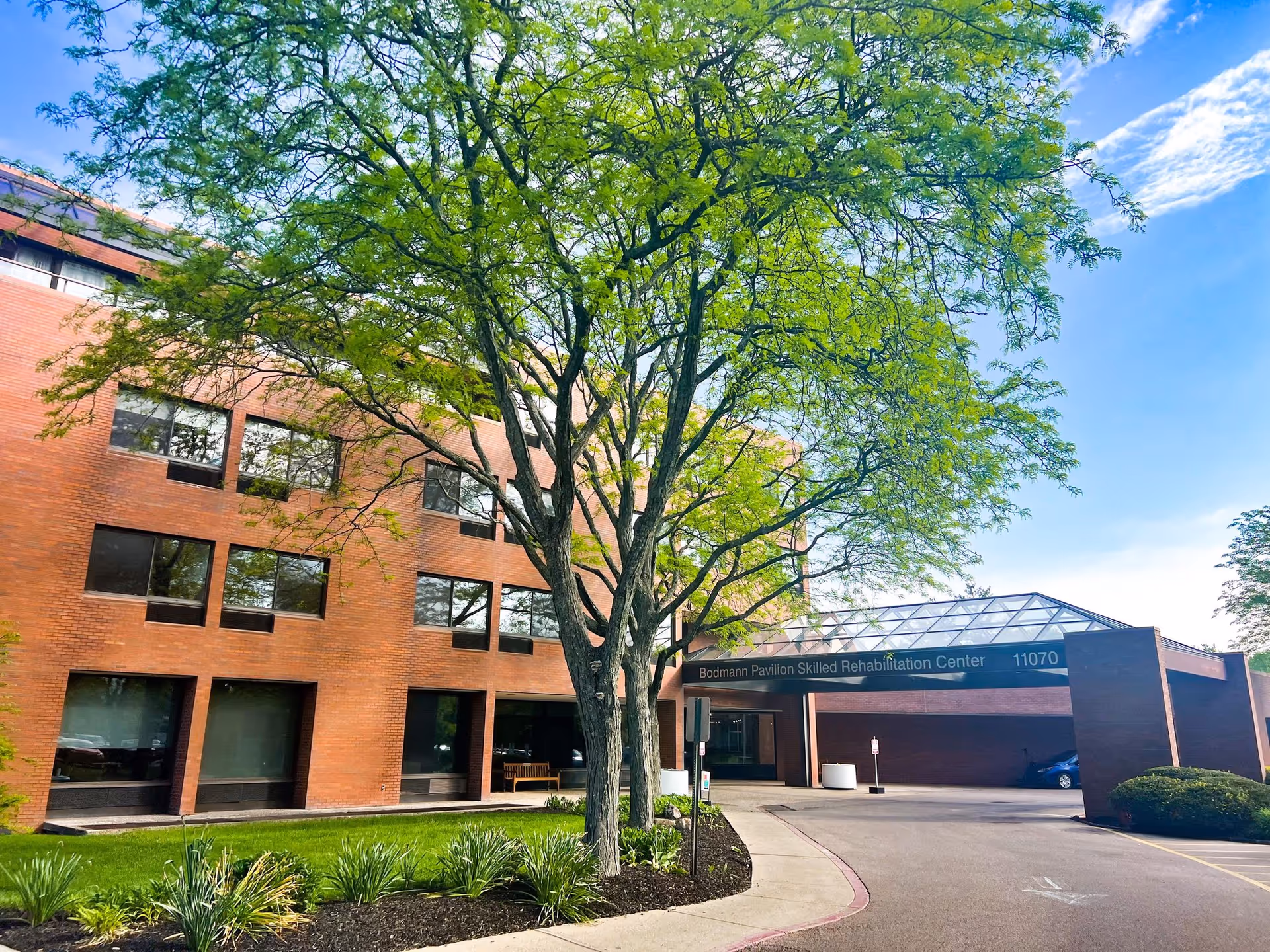 Brick facility front with glass entry canopy and green trees lining the walkway.