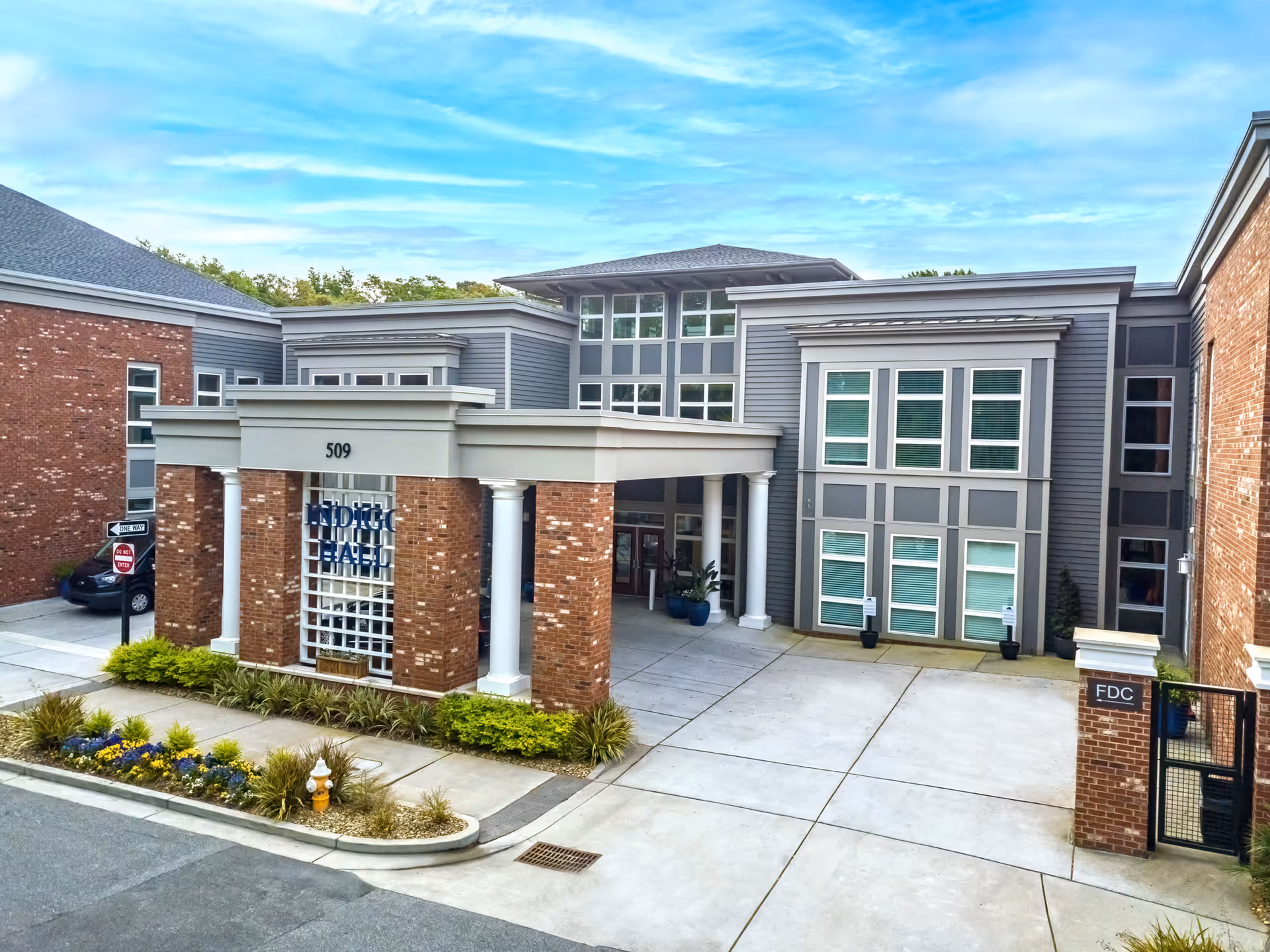 Front exterior view of Indigo Hall Assisted Living and Memory Care building with brick and gray siding, large windows, and a covered entrance supported by white columns. There are landscaped plants and flowers near the sidewalk and a fire hydrant in front.