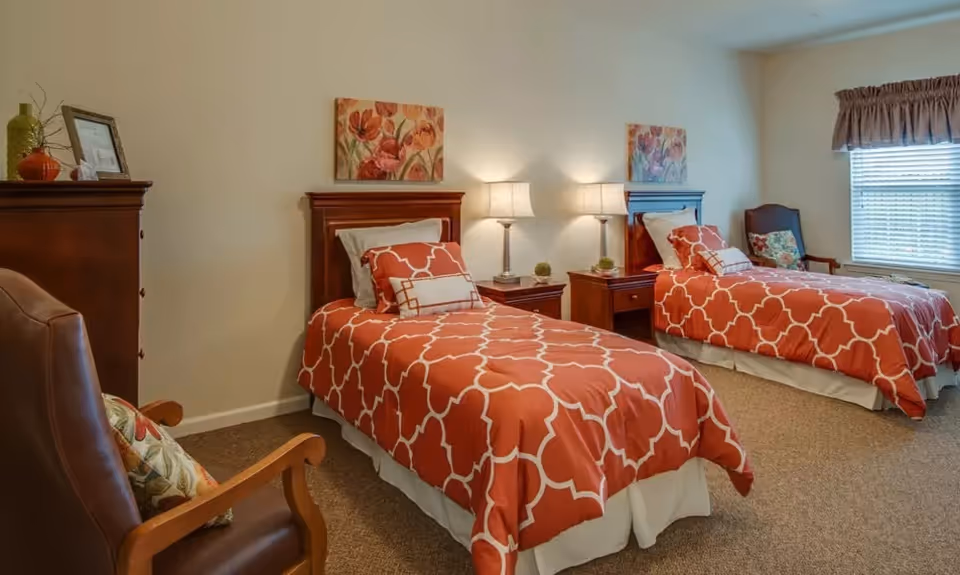 A senior living facility bedroom with two twin beds featuring orange and white patterned bedding. Each bed has a wooden headboard and is accompanied by a nightstand with a lamp. There are floral paintings above each bed, a window with blinds and a valance, and a wooden armchair with a floral cushion in the corner.