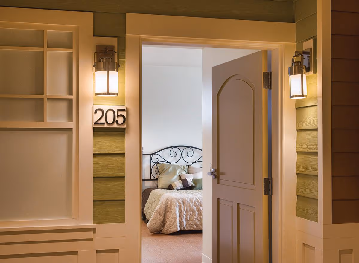 Open door reveals a bedroom with a metal bed frame, pillows, and warm lighting.