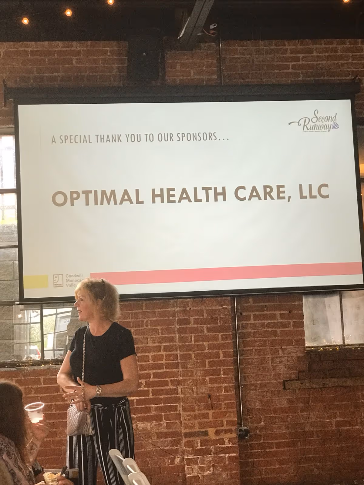 A woman stands beside a large screen displaying Optimal Health Care, LLC at an indoor event.