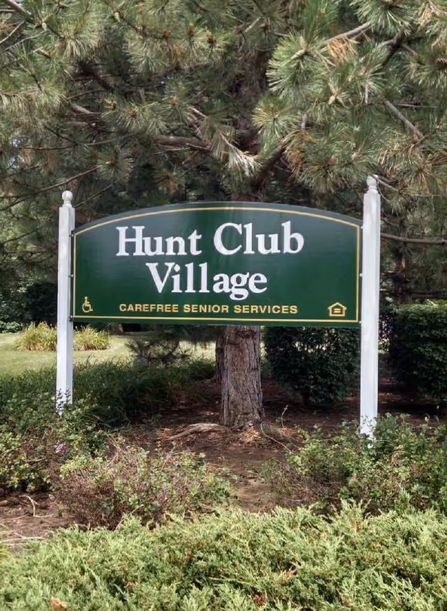 Green entrance sign reading "Hunt Club Village - Carefree Senior Services" set among trees and shrubs.