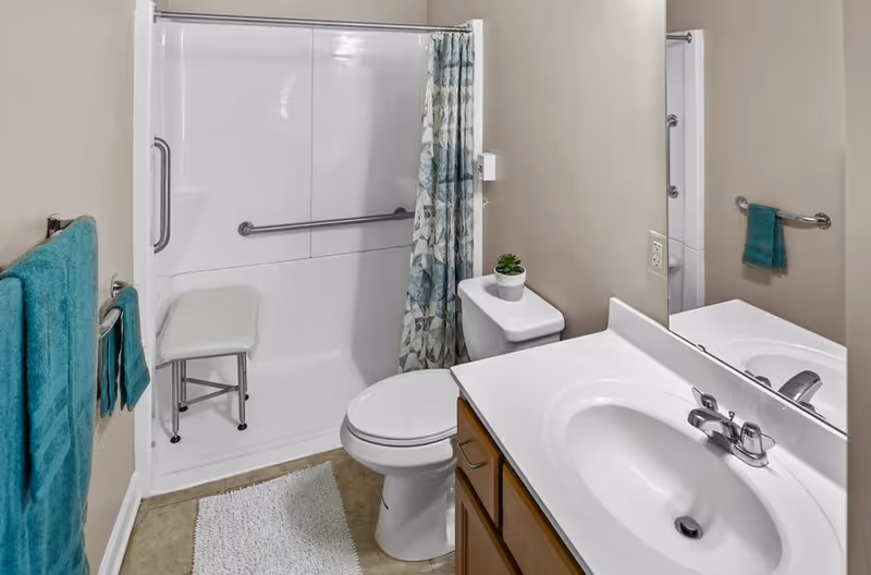Accessible bathroom with shower chair, toilet, sink, and teal towels.