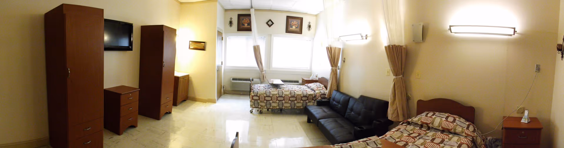 Panoramic view of a shared senior living bedroom with two beds, a black couch, wardrobe, wall-mounted TV, and windows.