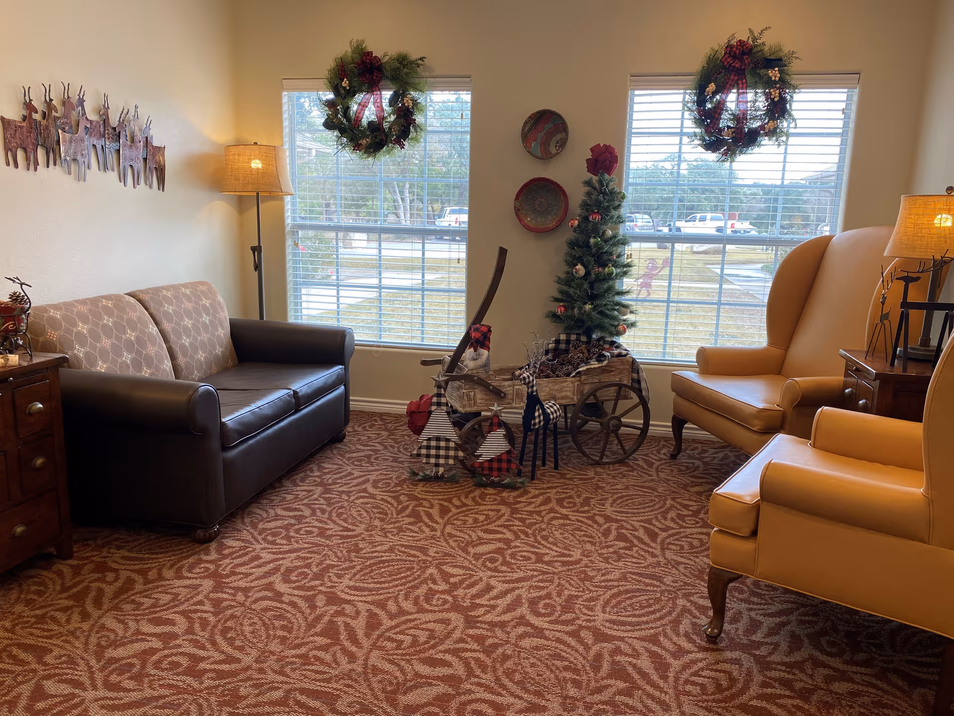 A cozy living room area with a patterned carpet, a brown and beige sofa on the left, two tan leather armchairs on the right, and a small decorated Christmas tree in a wooden cart between two large windows adorned with holiday wreaths. There are floor lamps on either side and wall decorations including metal reindeer and decorative plates.