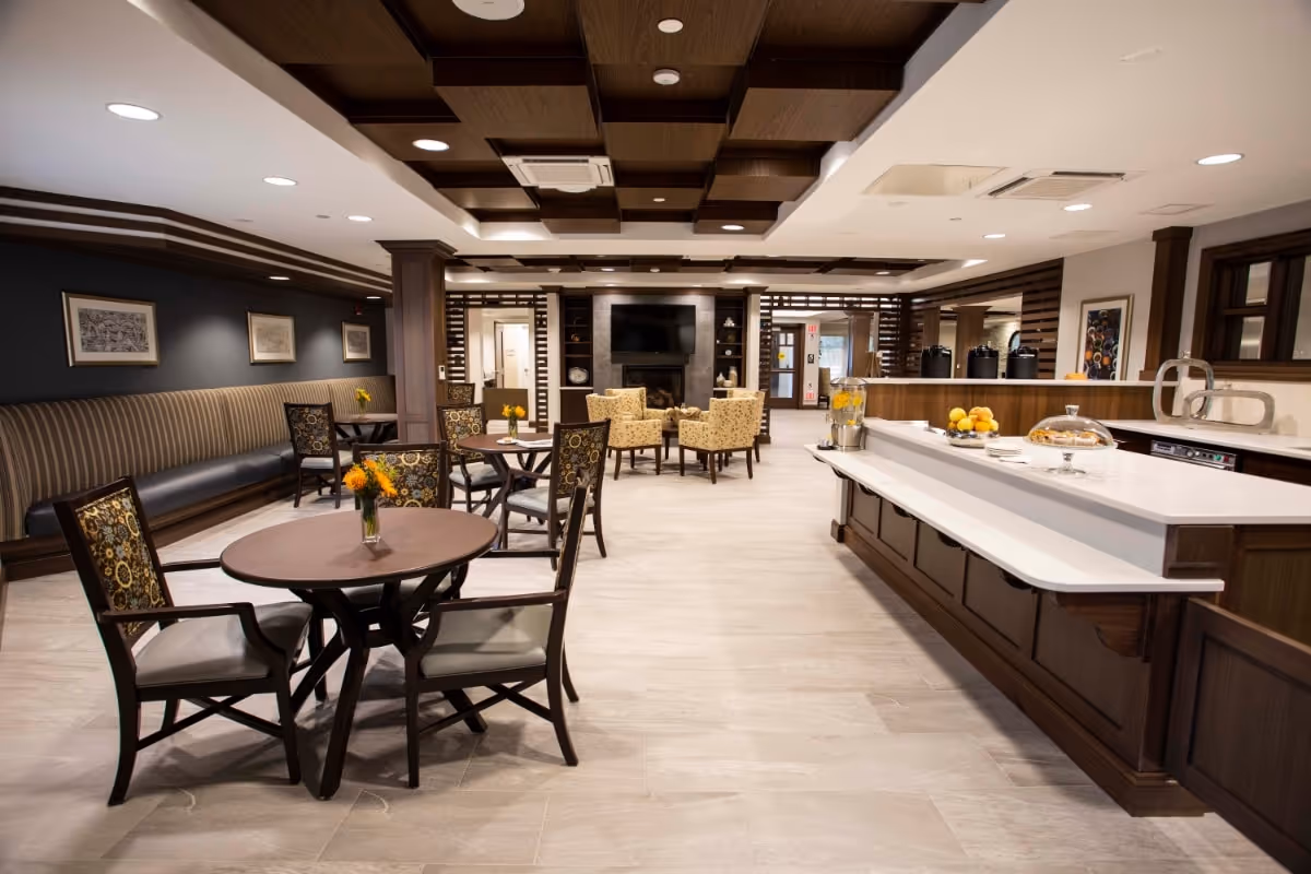 A spacious and well-lit common area in an assisted living facility featuring several round tables with chairs, a long cushioned bench along one wall, a counter with refreshments including a water dispenser and fruit, and a seating area with armchairs in front of a wall-mounted TV and fireplace.