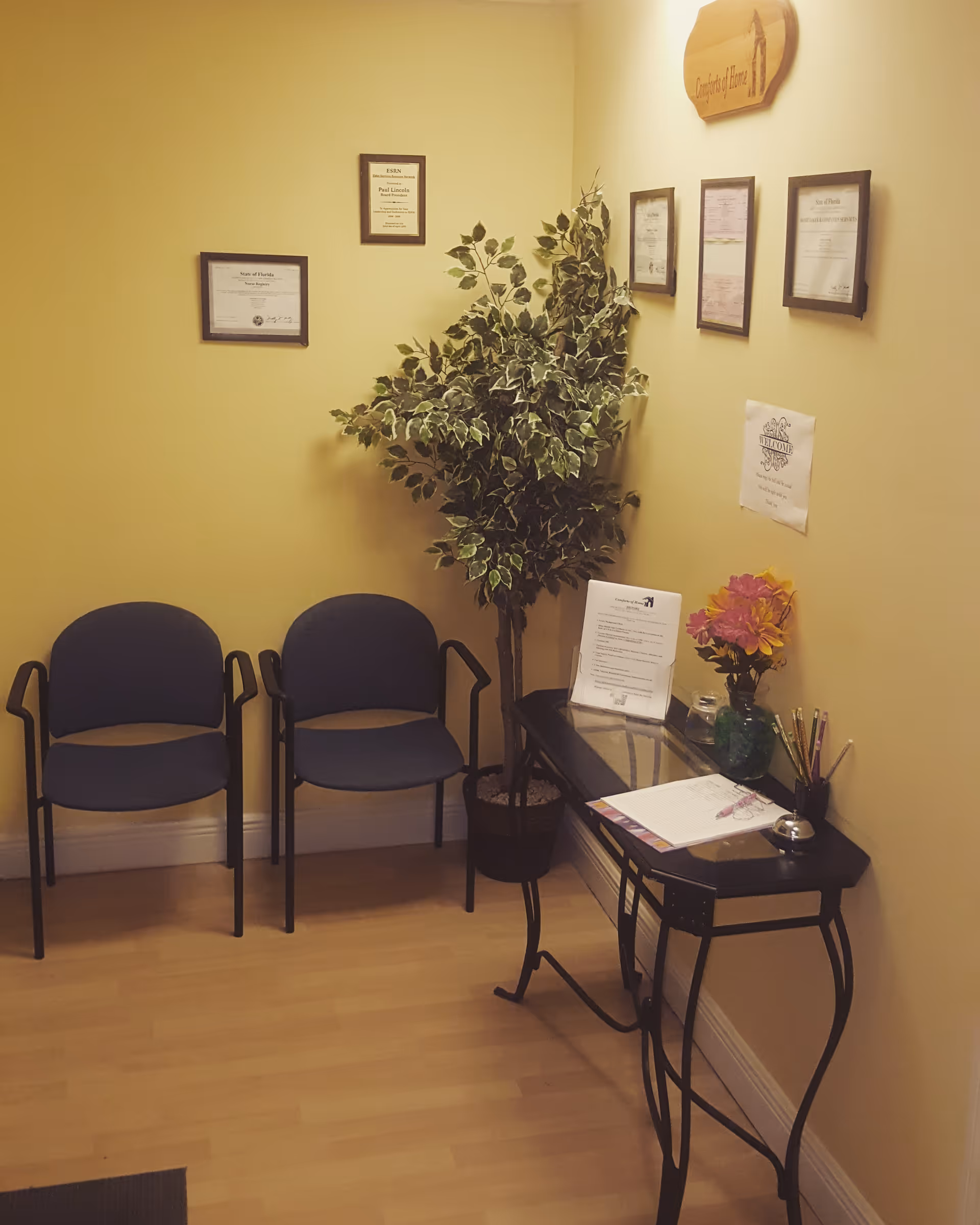 Waiting area with two chairs, a plant, and framed certificates on yellow walls.