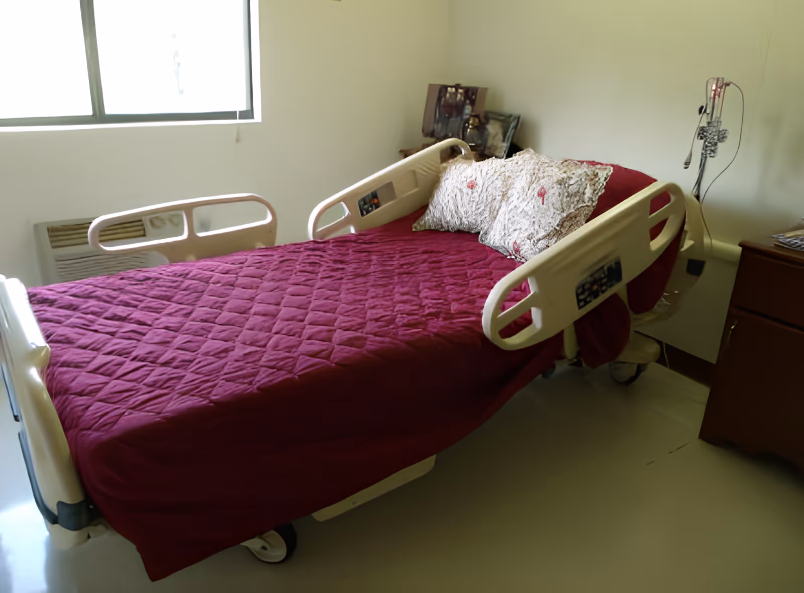 A hospital-style bed with a burgundy quilt and patterned pillows in a simple room next to a window and a bedside cabinet.