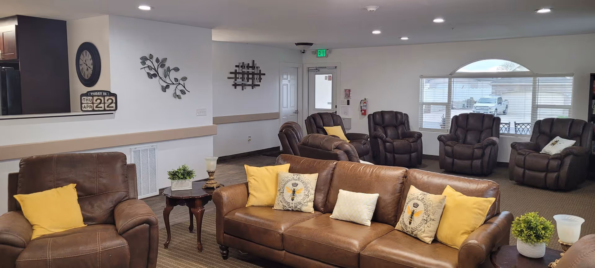 A spacious living room area with brown leather sofas and recliners arranged around the room. The sofas have yellow and patterned throw pillows. There are small wooden side tables with decorative plants and lamps. A large window with blinds lets in natural light, and the walls are decorated with a clock, a calendar showing Thursday April 22, and metal wall art. The room has a neutral color scheme with beige walls and carpeted floors.