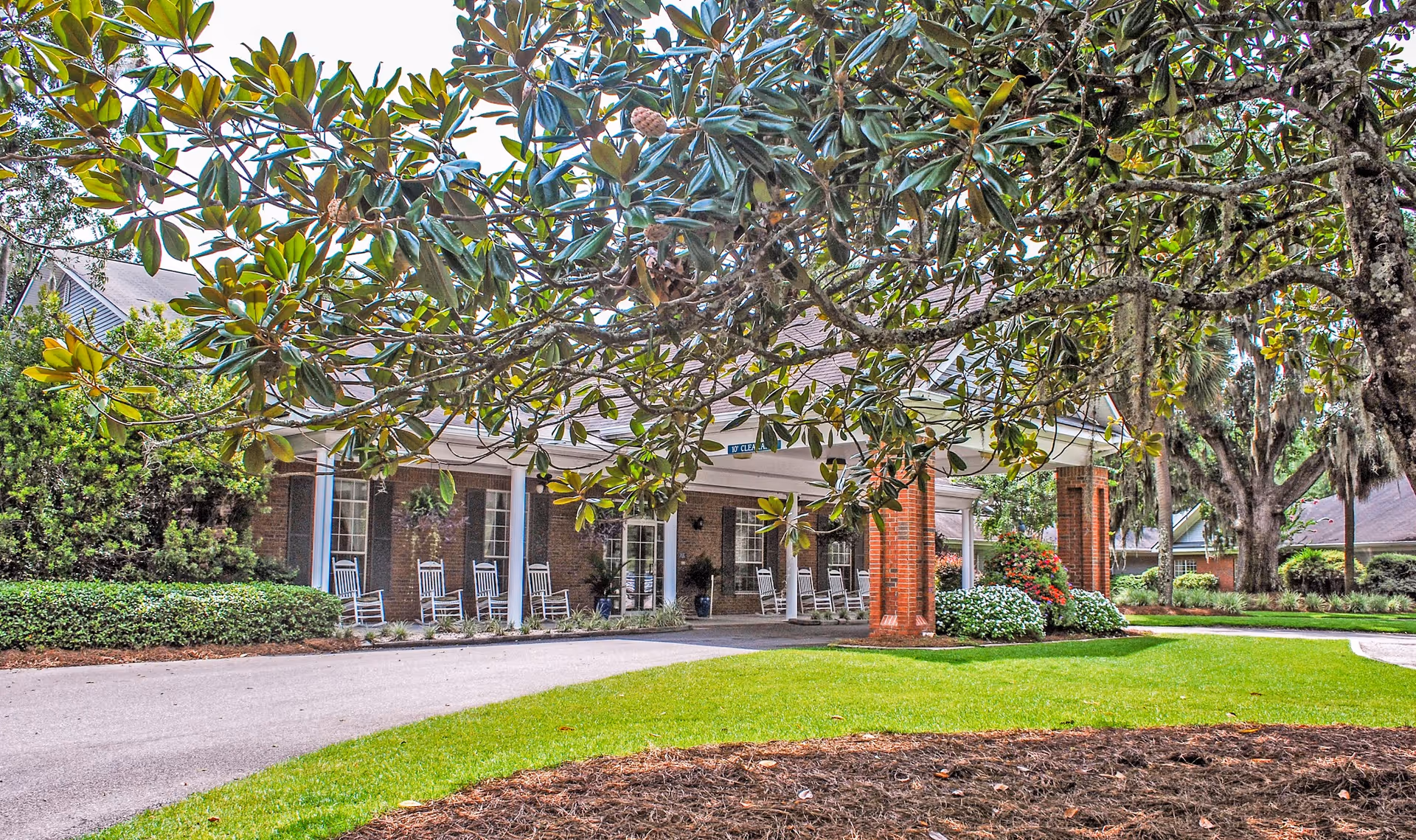 Front exterior view of a senior living facility with a covered entrance supported by brick columns. Several white rocking chairs are placed along the porch. The area is surrounded by well-maintained green lawns, bushes, and large trees with broad leaves.