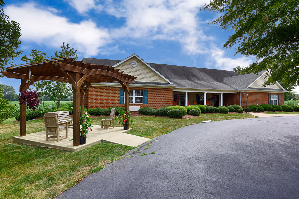 Exterior view of a single-story brick building with a gray roof, surrounded by neatly trimmed bushes and trees. In the foreground, there is a wooden pergola with hanging plants and outdoor seating on a concrete patio. The sky is partly cloudy with blue patches.