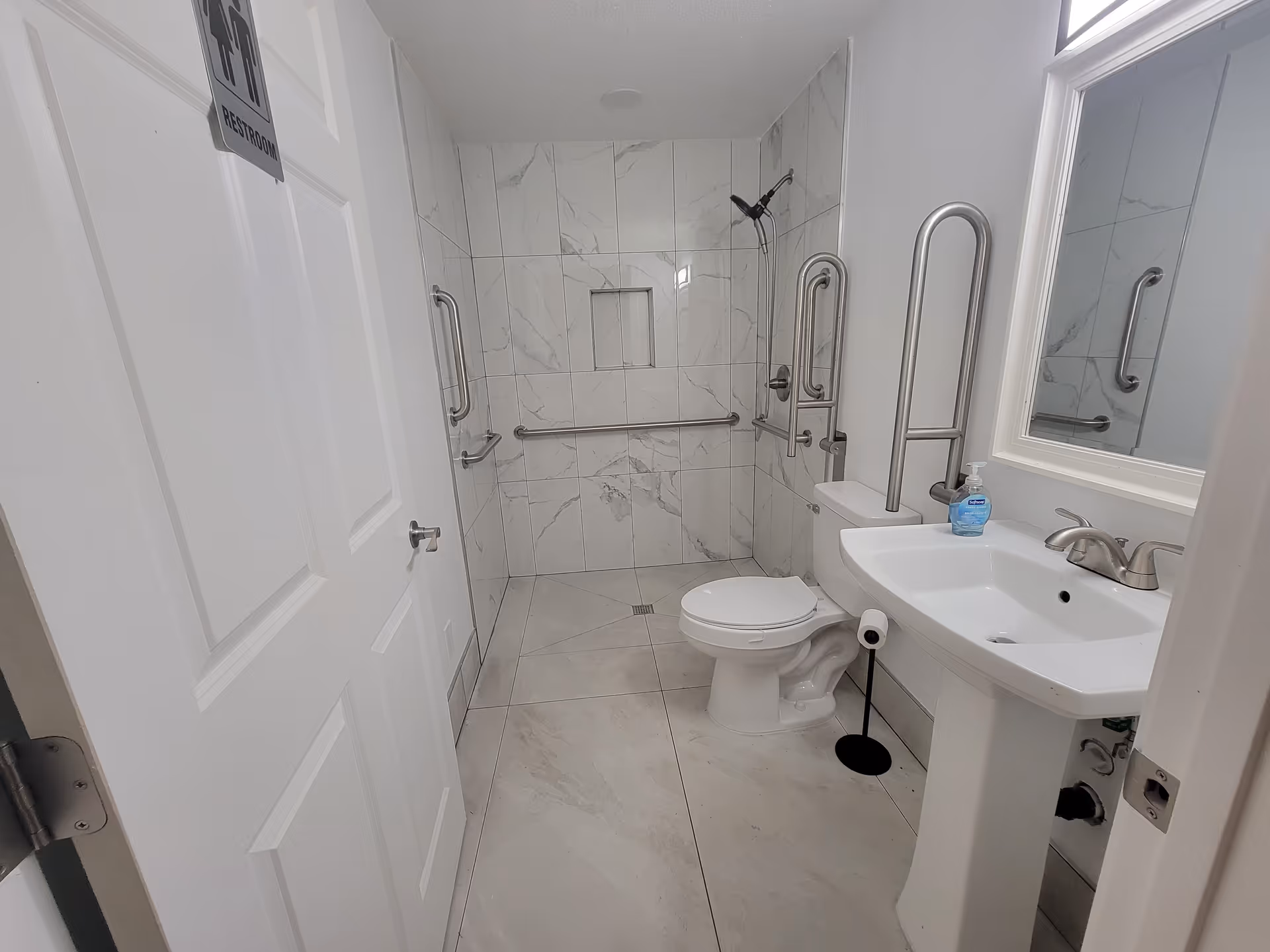 Accessible bathroom with white marble tiles, grab bars, toilet, and pedestal sink.