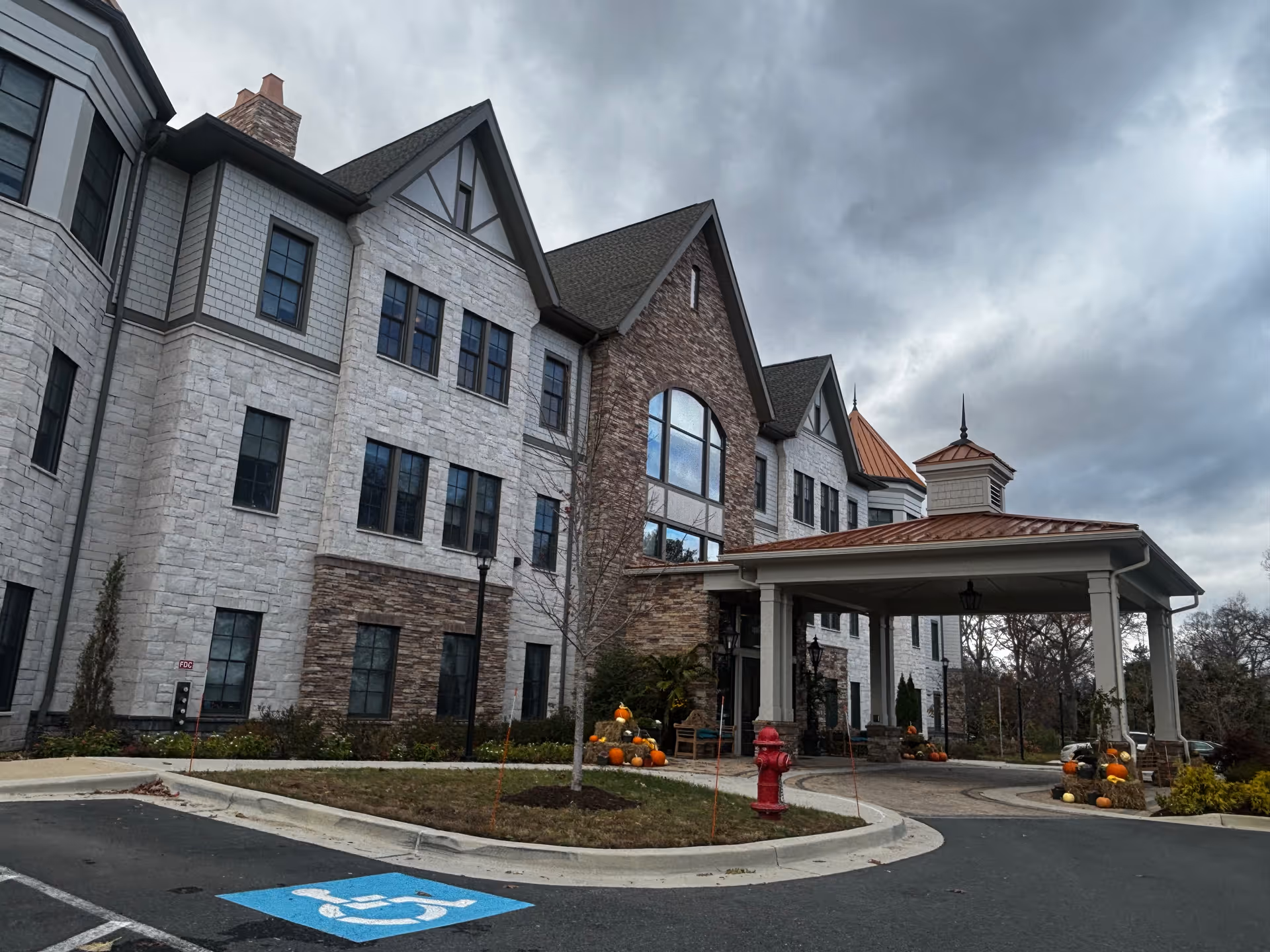 Front exterior of a multi-story stone and brick senior living building with a covered drop-off entrance, pumpkins on display, and a cloudy sky.