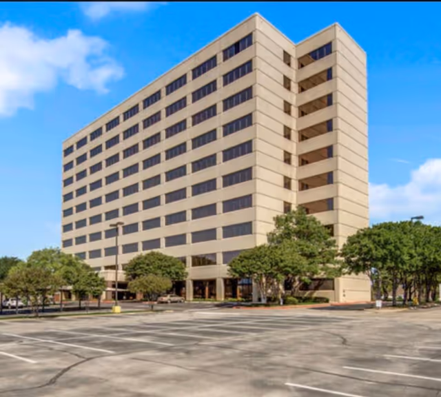 A tall beige multi-story building with a parking lot and trees in front.