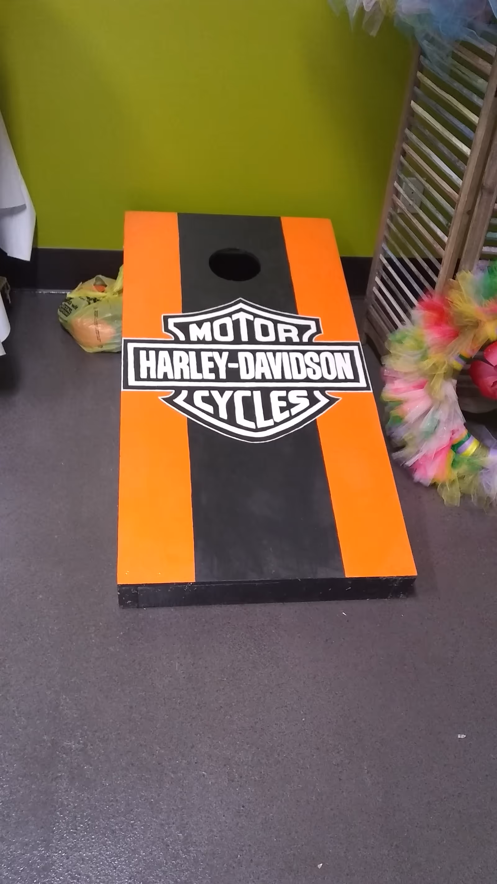 An orange and black Harley-Davidson cornhole board indoors against a lime wall.