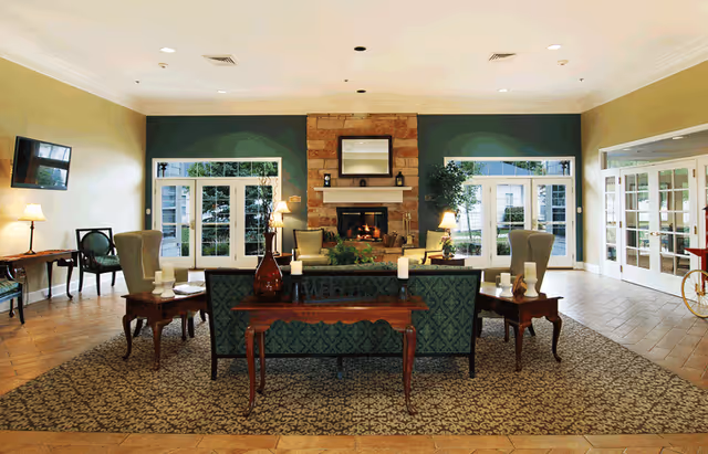 A spacious senior living facility common area with a patterned carpet, green upholstered sofas, wooden side tables with candles, and a stone fireplace in the center. Large windows and glass doors let in natural light, and there are several lamps and chairs arranged around the room.