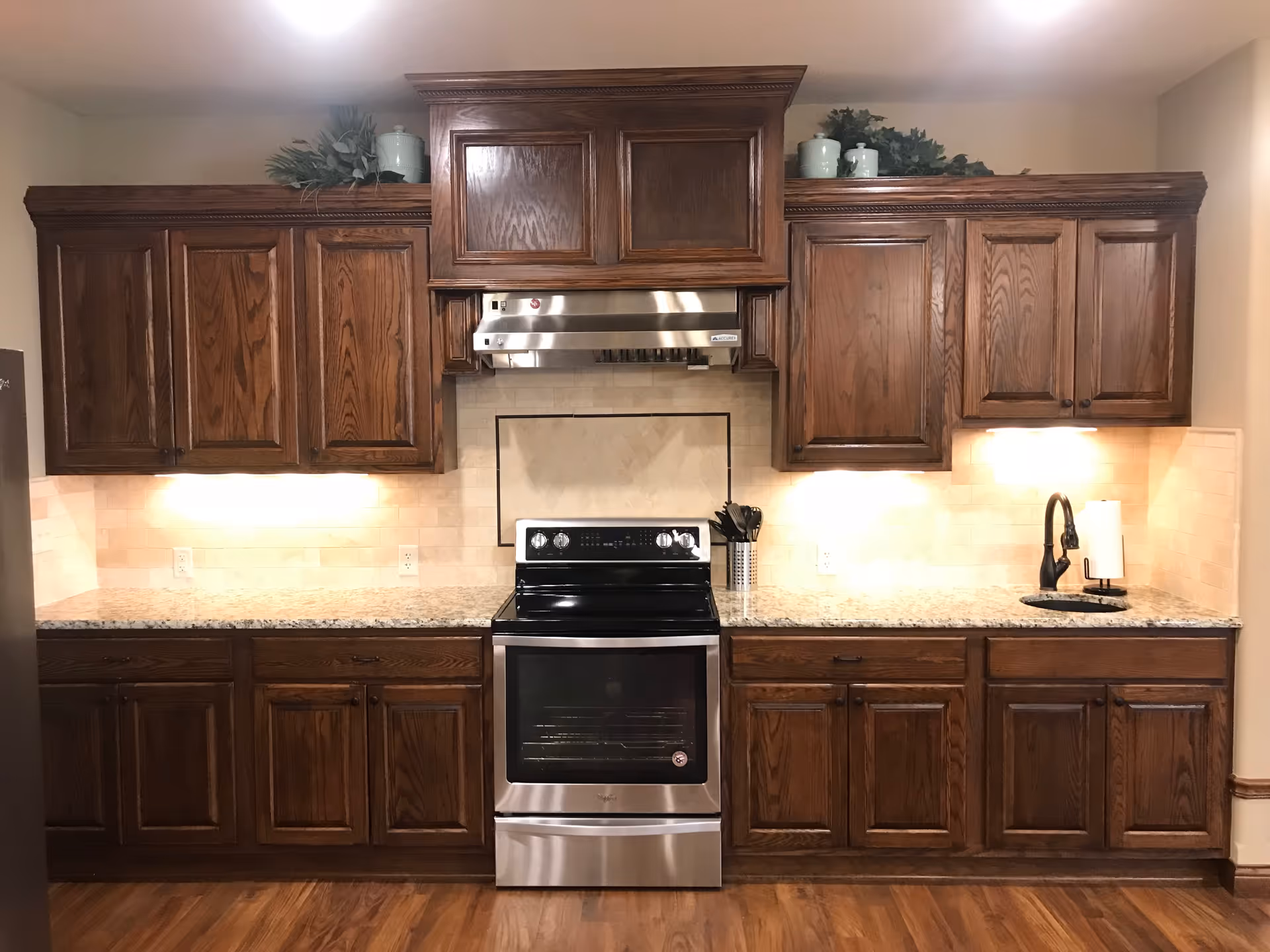 A kitchen with dark wooden cabinets, a stainless steel electric stove and oven, a stainless steel range hood, granite countertops, a small sink with a black faucet, and under-cabinet lighting. There are decorative plants and containers on top of the cabinets.