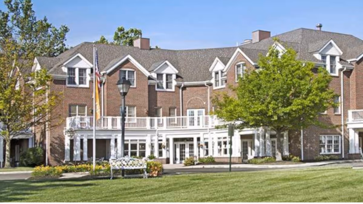 Front exterior of a multi-story brick senior living building with white porch, flagpoles, trees, and a bench on a lawn.