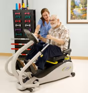An elderly man using a seated exercise machine with the assistance of a female caregiver in a room with light-colored walls, a painting on the wall, and a rack holding colorful dumbbells in the background.
