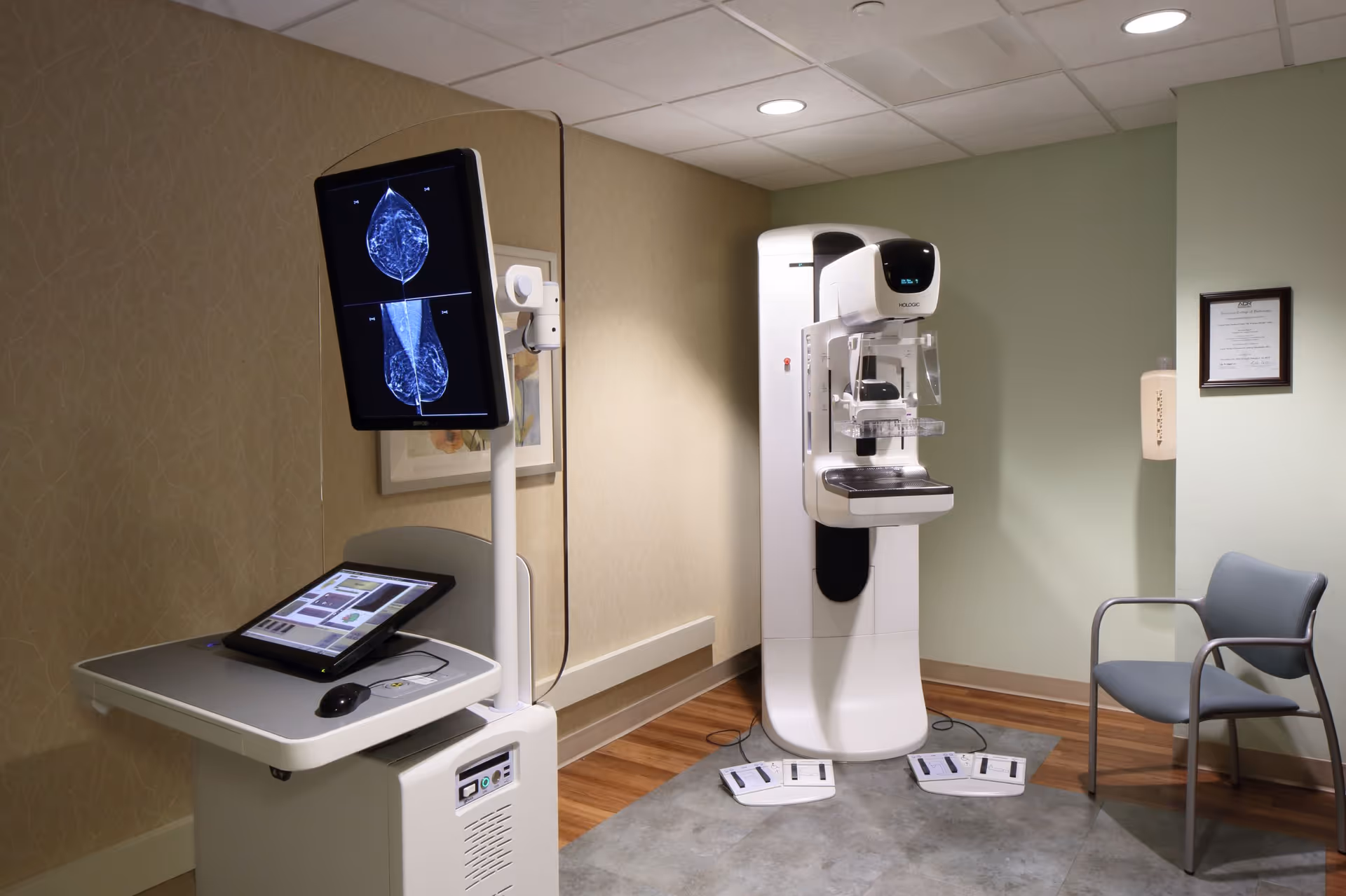 Clinical room with a mammography machine, computer console, and a waiting chair.
