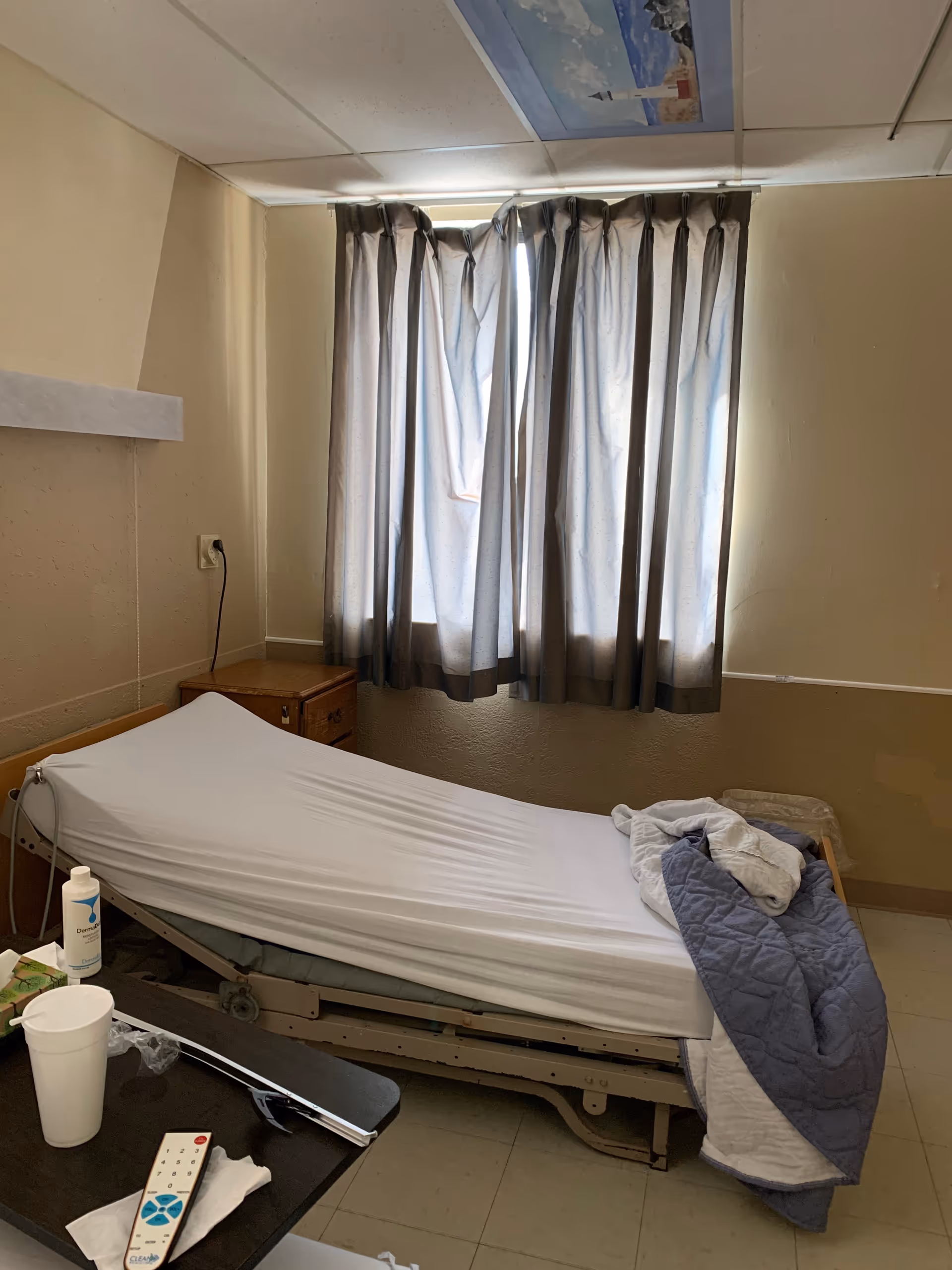 Hospital bed in a small bedroom with a window and gray curtains.
