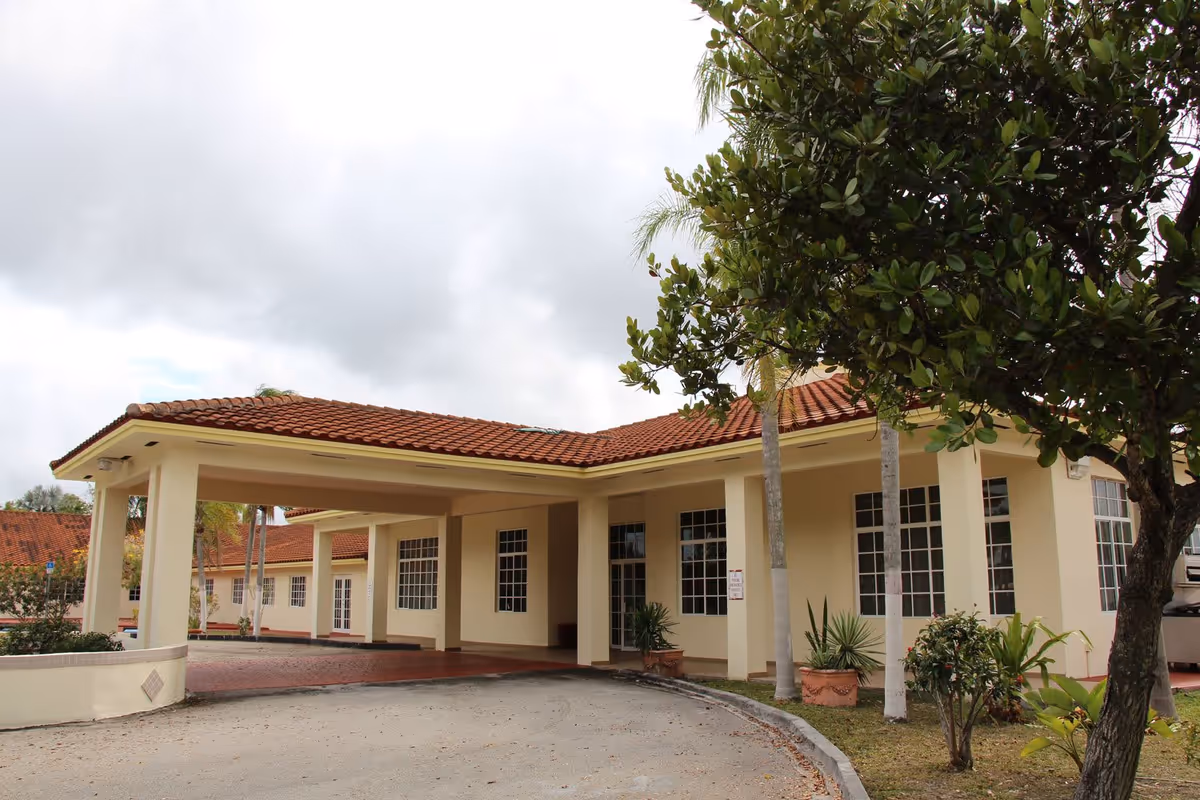 Entrance and covered porte-cochère of a single-story building with a red tile roof, large windows, and trees in front.