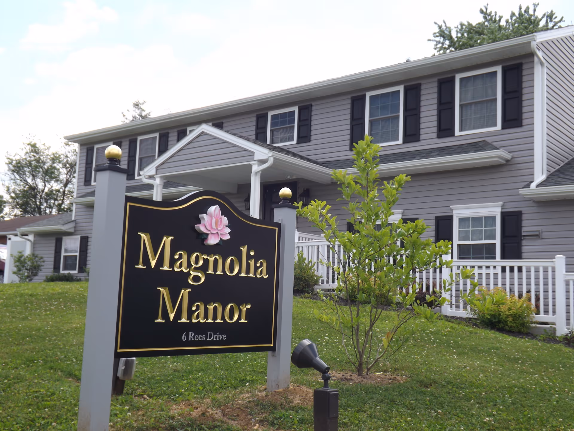 A black sign reading "Magnolia Manor" stands in front of a two-story gray building with white trim and a small lawn.