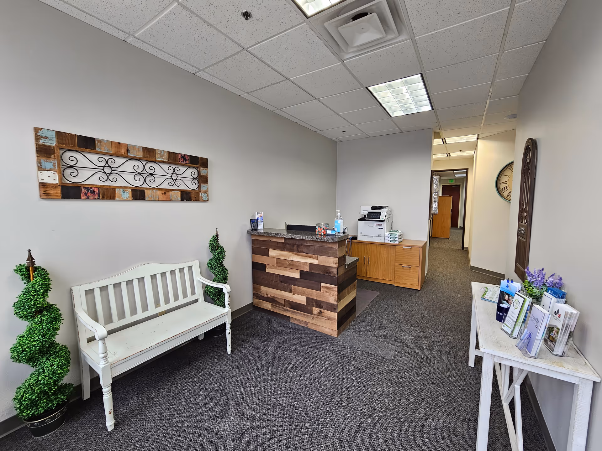 Reception area with a wooden front desk, white bench, and pamphlet table indoors.