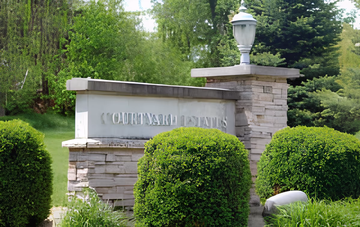 Stone entrance sign and pillar with a lamp surrounded by trimmed shrubs and trees.