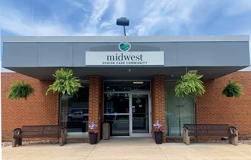 Front entrance of a brick senior care facility with glass doors, hanging plants, and benches.