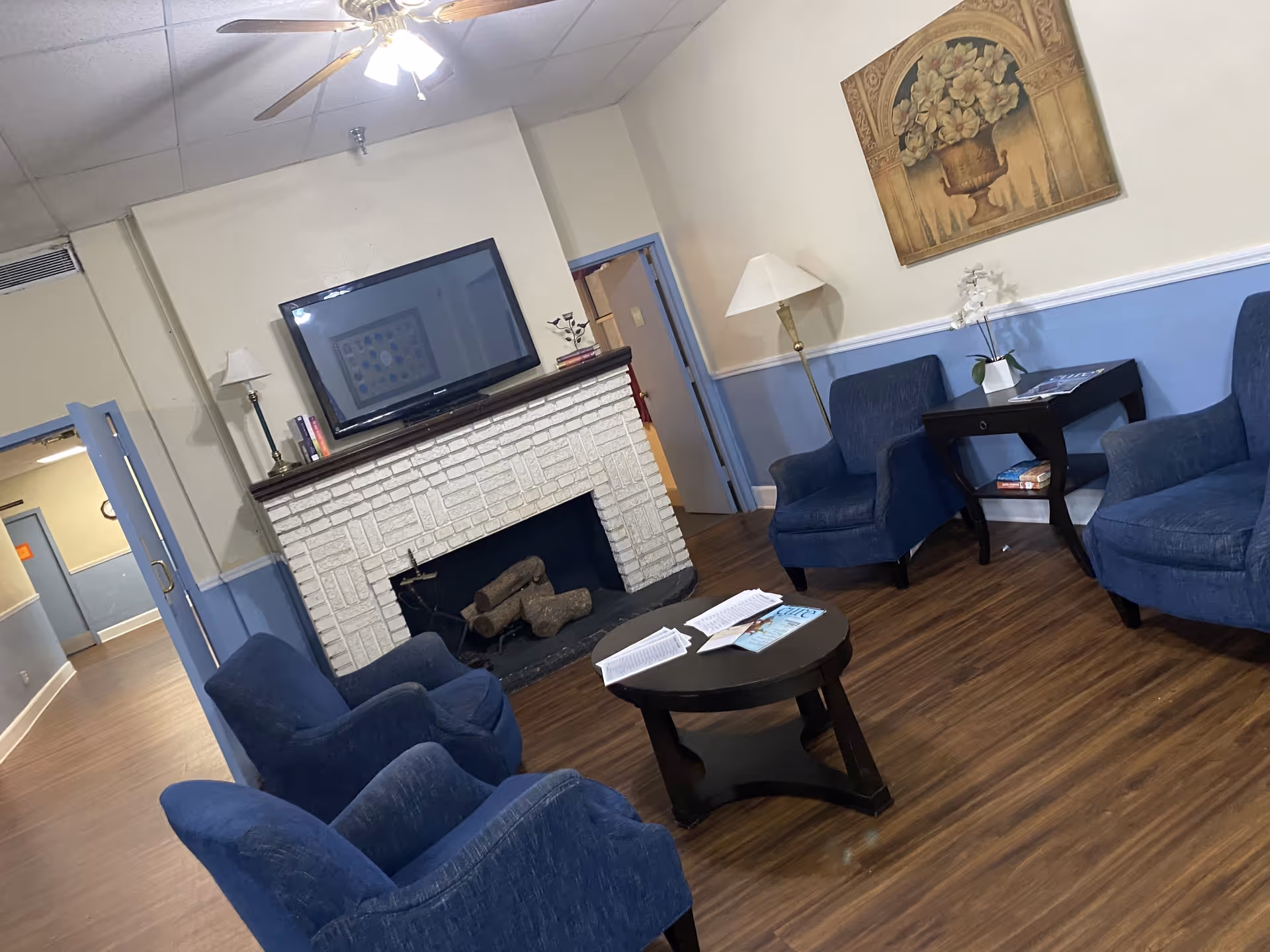 A cozy living room area with four blue upholstered armchairs arranged around a round wooden coffee table with papers and a book on it. There is a white brick fireplace with a flat-screen TV mounted above it. A floor lamp and a side table with a white orchid and books are placed near the chairs. The walls are painted beige on the upper half and blue on the lower half, with a floral painting hanging above the side table. The room has wooden flooring and a ceiling fan with lights.