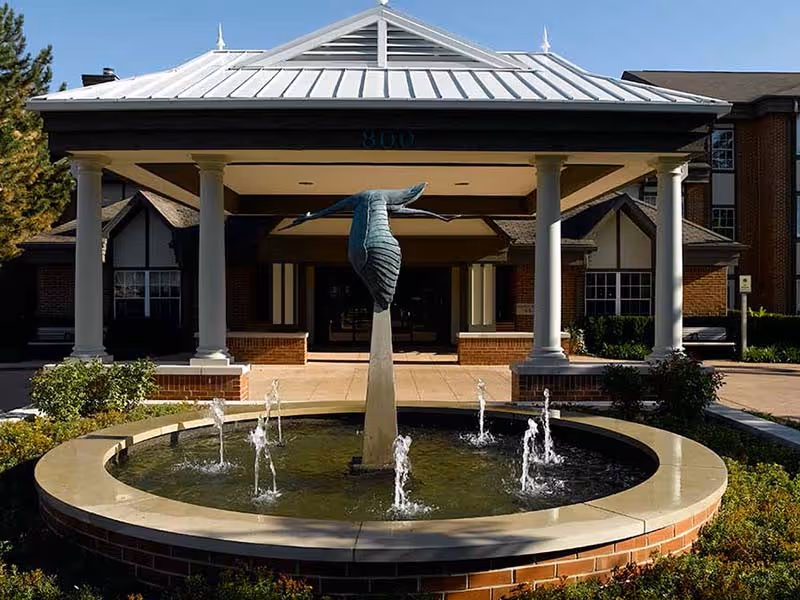 Entrance of a building with a covered portico supported by white columns, featuring a circular water fountain with a bird sculpture in the center, surrounded by greenery and brickwork.